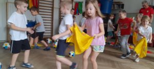 Dzieci w strojach gimnastycznych poruszają się trzymając w dłoniach żółte chusty 