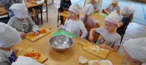 dzieci siedzą przy stolikach i kroją warzywa korzystając z deski i noża. Na środku stolika stoi metalowa miska 