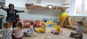 Dzieci siedzą przed nauczycielką i wykonują ruchy pokazane przez nią