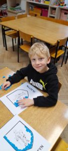 Chłopiec siedzi przy stoliku, przed nim rysunek herbu Olsztyna oraz plastelina 
