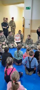 Sześcioro dzieci siedzi naprzeciw kilku osób w mundurach wojskowych, jedna kobieta trzyma godło Polski. 
