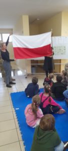 Kilkoro dzieci siedzi na materacach, patrząc w stronę flagi Polski trzymaną przez dwie osoby 