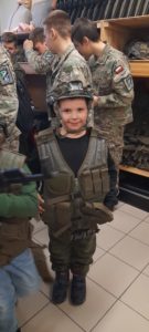 Chłopiec stoi w kamizelce kuloodpornej i hełmie, w tle osoby w mundurach wojskowych 