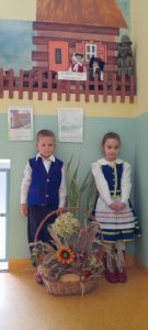 Chłopiec i dziewczynka stoją w strojach warmińskich na tle warmińskiej chaty. Przed nimi kosz z jesienną kompozycją 