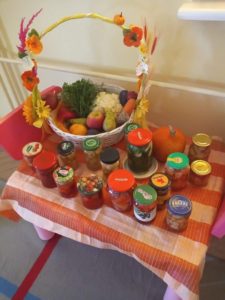 Na stoliku z lewej strony stoi kosz z owocami i warzywami. Na pozostałej części stolika umieszczone są słoiki z przetworami. 