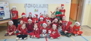 Grupa przedszkolaków ubranych na czerwono z czapkami Mikołaja, w tle na tablicy magnetycznej napis "Mikołajki" 