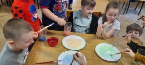 Dzieci siedzą przy stoliku. Moczą pędzle z farbą w mleku. 