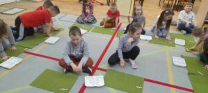 Dzieci siedzą na dywanie, przed nimi leżą zielone podkładki na których ułożony jest groch. Obok podkładek znajdują się białe plastikowe pudełka z grochem 