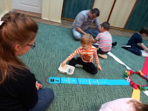 Dwoje rodziców i troje dzieci siedzą na dywanie i układają rytmy z wykorzystaniem patyczków i fasolek
