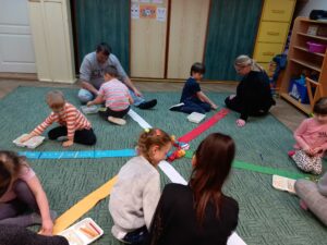 3 rodziców oraz 6 dzieci siedzą na dywanie i układają rytmy z patyczków i fasolek na paskach wiatraka matematycznego