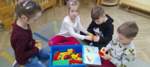 Na dywanie siedzi czworo dzieci (dwie dziewczynki i dwóch chłopców), przed nimi pudełko z klockami Lego oraz instrukcja