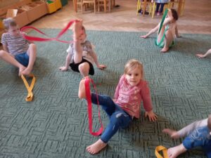 Czworo dzieci wykonuje ćwiczenie gimnastyczne - próbują w palcach stóp utrzymać szarfę