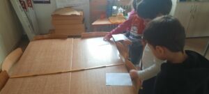 Troje dzieci stoi przy stoliku i mierzy jego szerokość za pomocą miary stolarskiej 