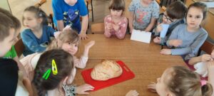 Kilkoro dzieci siedzi przy stoliku na którym znajduje się deska do krojenia a na niej Chleb 
