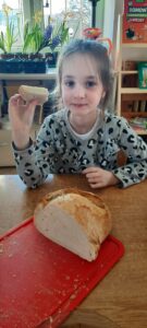 Dziewczynka siedzi przy stoliku, w dłoni trzyma kromkę chleba, przed nią na desce do krojenia znajduje się chleb 
