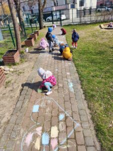Dzieci rysujące kredą na chodniku w ogrodzie przedszkolnym.