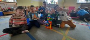 sześcioro dzieci siedzi na dywanie. Jedno z nich trzyma obrazek Wysokiej Bramy. Przed nimi stoi Wysoka Brama zbudowana z klocków Lego. 