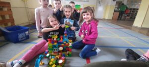 Kilkoro dzieci siedzi na dywanie. Jedno z nich trzyma obrazek Zamku Kapituły Warmińskiej. Przed dziećmi znajduje się Zamek zbudowany z klocków Lego. 