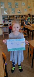 Dziewczynka stoi, trzyma w dłoniach kartkę z wykonanym rysunkiem