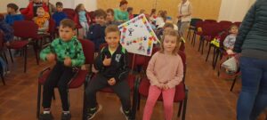 Na pierwszym planie troje dzieci siedzi na krzesłach, między nimi logo przedszkola. W tle kilkoro dzieci oraz krzesła. 