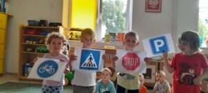 Czworo dzieci w sali przedszkolnej stojąc trzyma obrazki przedstawiające znaki drogowe: przejście dla pieszych, Stop, parking, droga dla rowerów. W tle widać dwoje siedzących dzieci