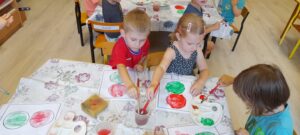 Troje dzieci siedzi przy stoliku i maluje farbami szablon sygnalizatora świetlnego .