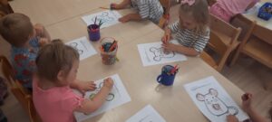 Dzieci siedzą przy stoliku I kredkami kolorują obrazek myszki 