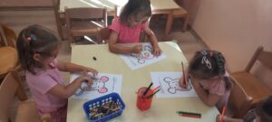 Trzy dziewczynki siedzą przy stoliku I kredkami kolorują obrazek myszki