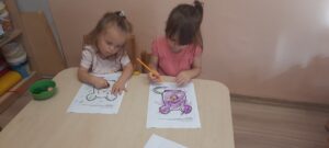 Dwie dziewczynki siedzą przy stoliku I kredkami kolorują obrazek myszki