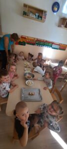 Grupa dzieci siedzi przy stoliku. Na środku stolika znajdują się miski z pokrojonymi owocami 
