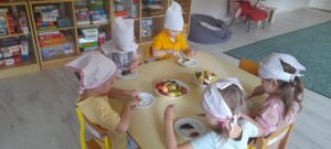 Pięcioro dzieci kroi owoce sezonowe siedząc przy stoliku . Na stoliku są talerze z owocami.