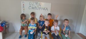 8 chłopców siedzi na krzesłach, trzymając medale i lizaki. W tle na białej tablicy wisi napis DZIEŃ CHŁOPAKA