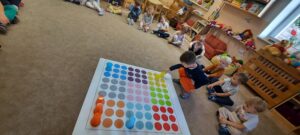 Dzieci siedzą na dywanie, na środku znajduje się mata do kodowania i kolorowe kubeczki. Chłopiec ustawia kolorowy kubek na macie. 