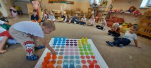 Dzieci siedzą na dywanie, na środku znajduje się mata do kodowania i kolorowe kubeczki. Chłopiec ustawia kolorowy kubek na macie. 