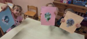 Troje dzieci siedzi przy stoliku, w dłoniach trzymają kolorowe baloniki papierowe