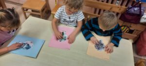 Troje dzieci siedzi przy stoliku, palcem naciskają kolorowe elementy znajdujące się na kartce 