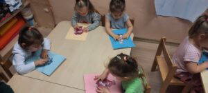 Czworo dzieci siedzi przy stoliku, palcem naciskają kolorowe elementy znajdujące się na kartce 