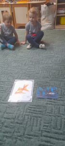 Na dywanie leżą ilustracje przedstawiające rybki. W tle 2 siedzących dzieci.