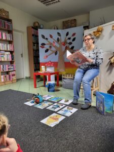 Pani Bibliotekarka czytająca książkę. Na dywanie obrazki przedstawiające niedźwiadki z bajek.