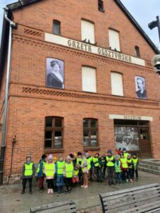 Grupa dzieci w odblaskowych kamizelkach w tle budynek z czerwonej cegły z napisem Gazeta Olsztyńska.