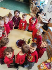 Grupa przedszkolaków ubrana na czerwono/bordowo stojąca przy stole na którym są buraki w misce, oliwa i przyprawy.