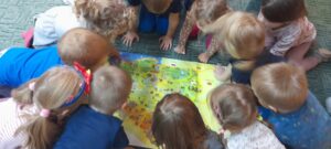 Na dywanie leży mapa z konturem Polski. Wokół mapy znajdują się dzieci, które ją oglądają, okreslają kształt, kolory, wskazują ważne miejsca.