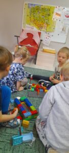 Na dywanie siedzą dzieci i budują zgodnie z poleceniem domy z klocków Lego.