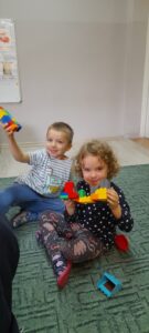 Na dywanie siedzi dwoje dzieci i prezentuje swoje skonstruowane domy z klocków Lego.
