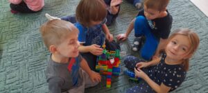 Na dywanie jest grupa dzieci , która prezentuje wspólnie zbudowany dom z klocków Lego.