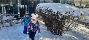 Dwoje dzieci stoi obok krzaka, na trawniku i krzakach jest śnieg 