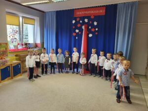 Dzieci ustawione w półkolu, recytują wiersz. W tle dekoracja w barwach Polski, z napisem : Narodowe Święto Niepodległości.