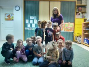 Grupa dzieci siedząca na dywanie Z tyłu nauczycielka. Jedno z dzieci trzyma maskotkę wiewiórkę.