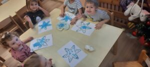 Grupa dzieci siedzi przy stoliku malując palcami śnieżynki niebieską farbą 