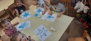 Kilkoro dzieci siedzi przy stoliku malując palcami śnieżynkę niebieską farbą 